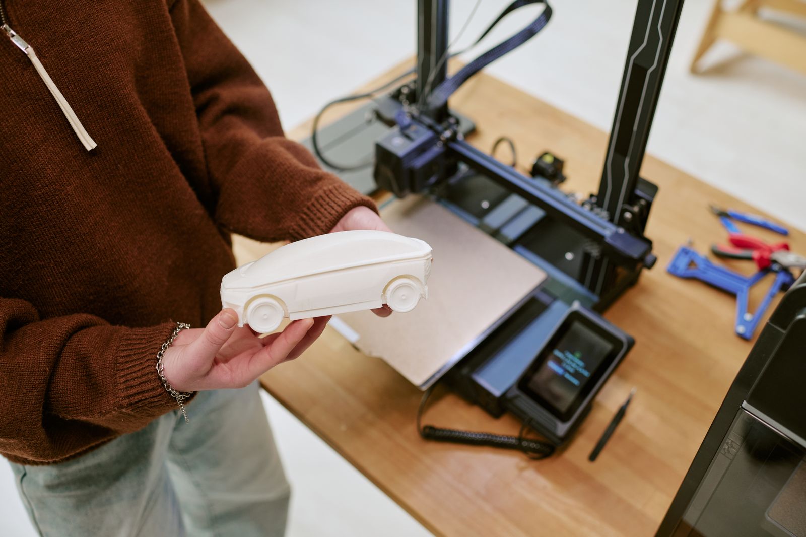 A person holding a 3D-printed prototype car model next to the printer that made it A person holding a 3D-printed prototype car model next to the printer that made it